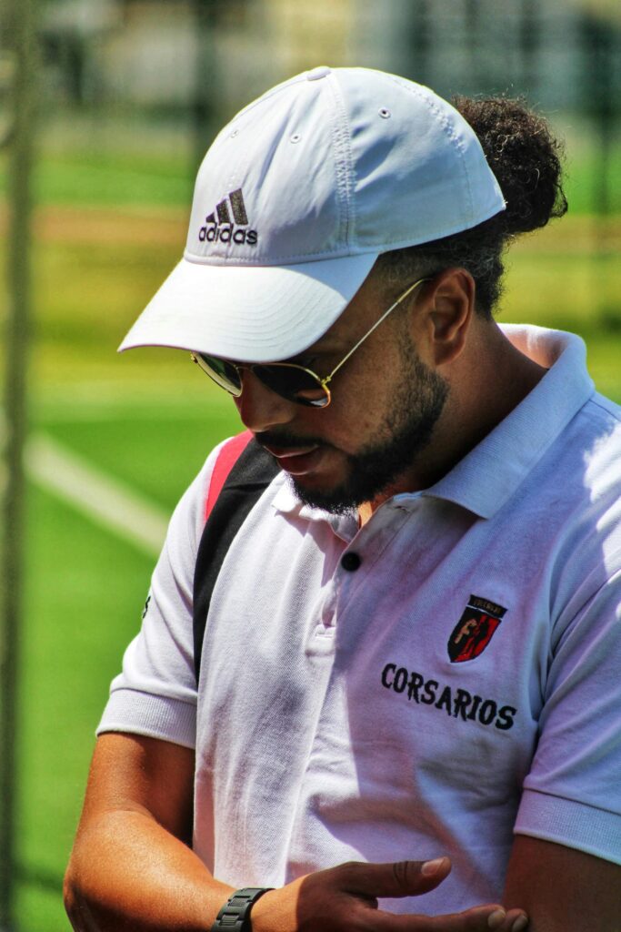 A male sports coach outdoors wearing a branded white cap and sunglasses on a sunny day.